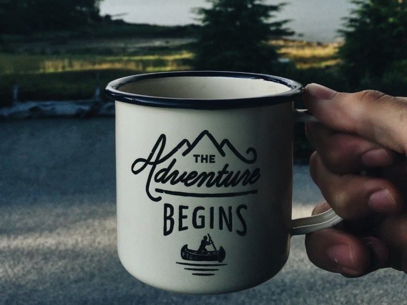 Hand holding mug that is imprinted 'The Adventure Begins'